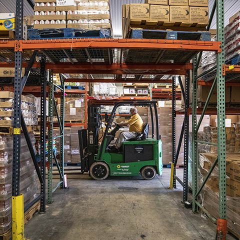 Man on a forklift in the Palm Beach County Food Bank's warehouse, which has shelves filled with boxes and cans of food