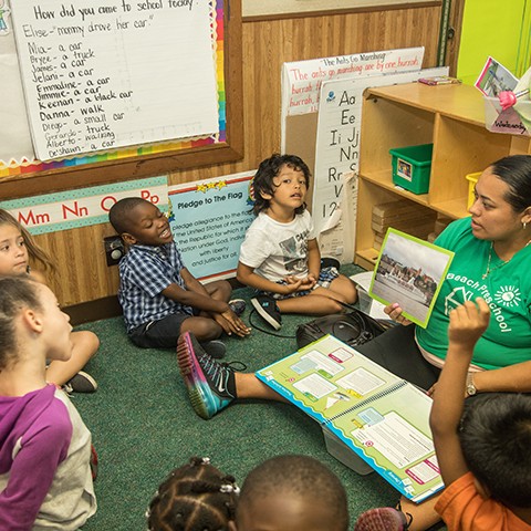 Preschool teacher in a classroom reads a book to young children sitting in a circle. One girl stands, one boy raises his hand.