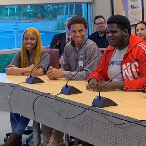 Three high school students from the Student Aces program smile while they sit at a conference table presenting their grant-making project to Children's Services Council's board. 