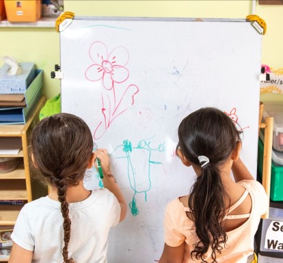 Two little girls stand in front of an easle drawin with colored markers in a child care center.