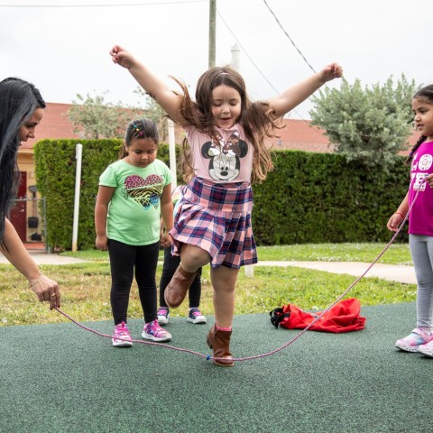 Three little girls play jump rope with a child care teacher on a playground.