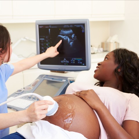 A pregnant woman with her partner getting an ultrasound, with a tech showing them the baby on the screen, in a doctor's office.