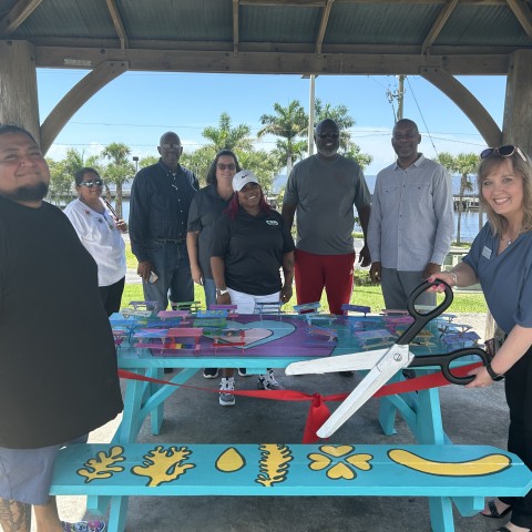 CSCPBC CEO Lisa Williams-Taylor, accompanied by teaching artist Jose Mendez, cuts a red ribbon with oversized scissors at the unveiling of the Pahokee Community Art Project in July 2024.