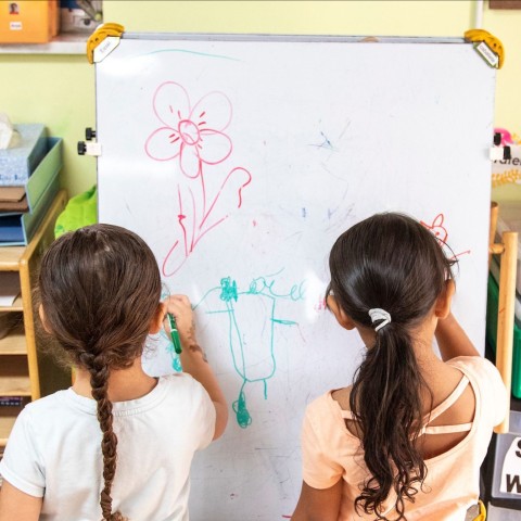 Two little girls stand in front of an easle drawin with colored markers in a child care center.
